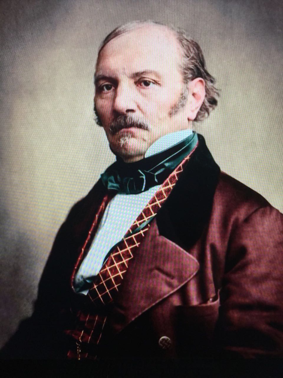 Portrait of a man with a serious expression, wearing a burgundy coat, geometric-patterned vest, and green cravat, against a neutral background.