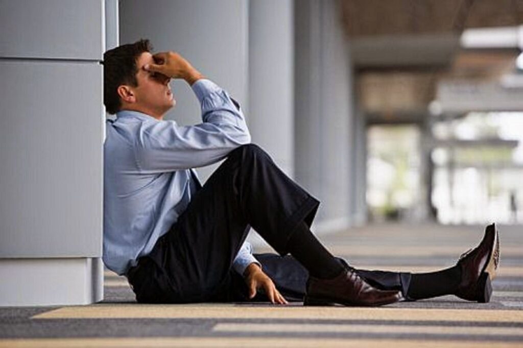 Man in a suit sitting on the floor against a wall, holding his face in a gesture of regret and distress.