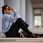 Man in a suit sitting on the floor against a wall, holding his face in a gesture of regret and distress.
